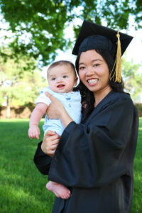 A young mother in a graduation cap and gown proudly holds her young child.